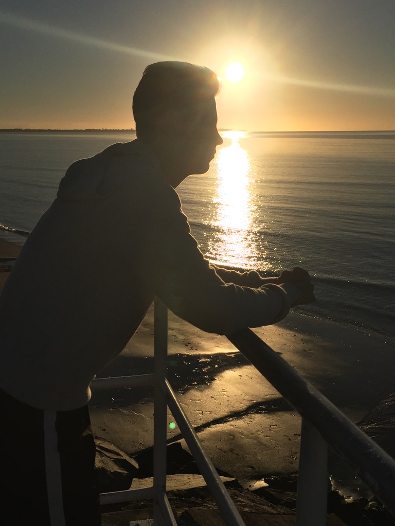Young person in silhouette at sunset by the water, leaning on a railing